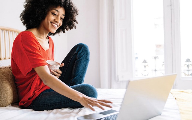 A woman sitting on her bed smiling while using her laptop.