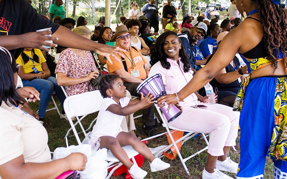 A woman hands a drum to a young girl sitting beside Ashley Brooks at Ruby's Happy Farm