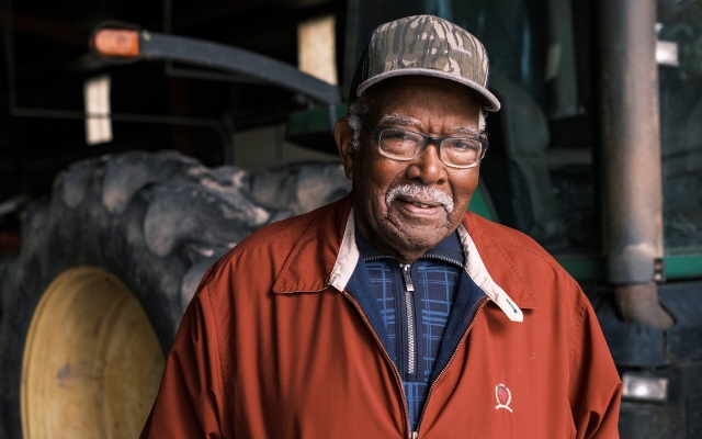 Man in an orange windbreaker stands in front of a green tractor.